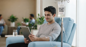 A smiling young man in a grey sweater seated in a light blue recliner, receiving an intravenous (IV) infusion while using a digital tablet. The IV bag is clearly labeled "REVITALIZE WELLNESS - HIGH-DOSE VITAMIN C INFUSION." A blurred waiting area with plants and other people is in the background.